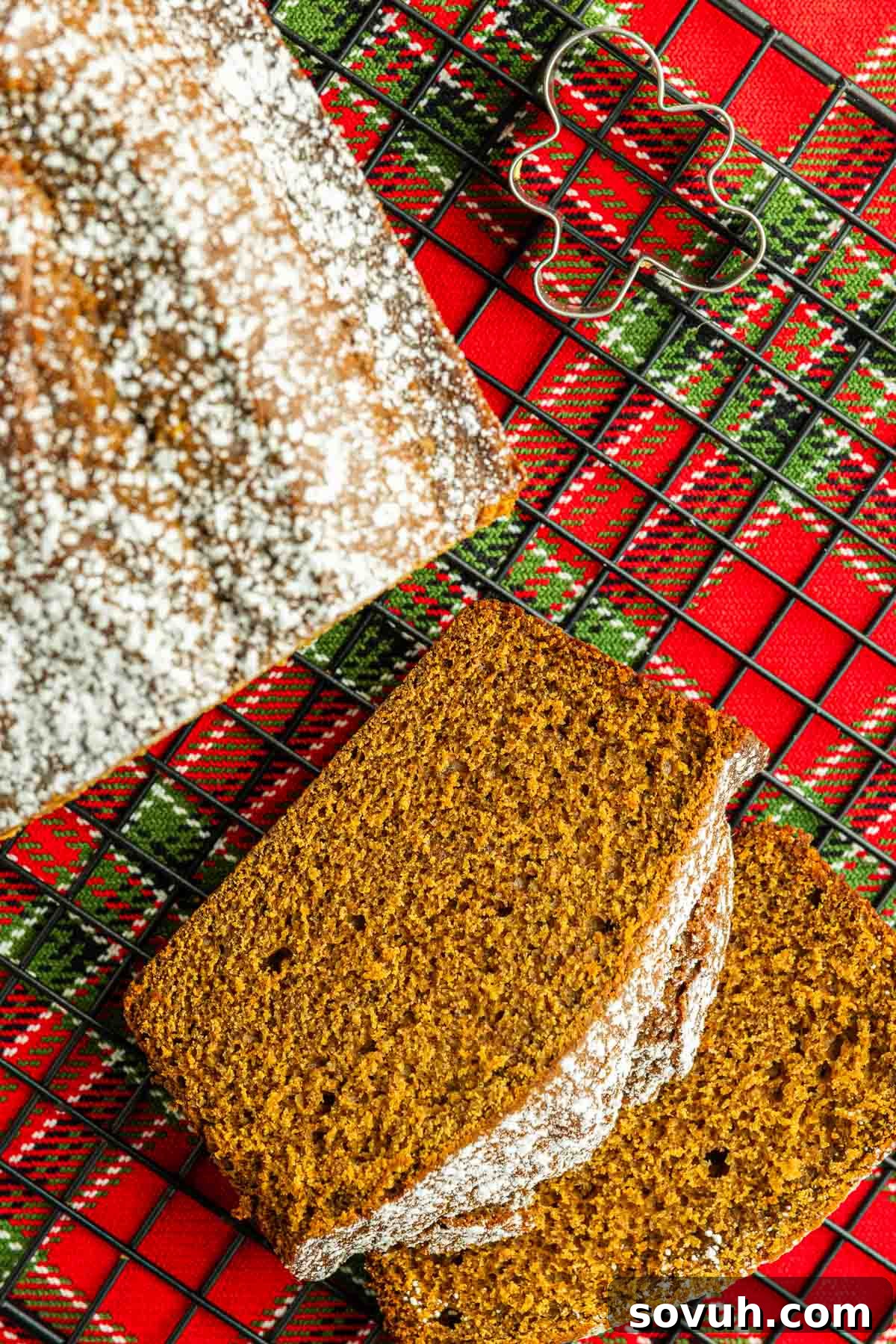 A beautifully sliced homemade gingerbread loaf, generously dusted with powdered sugar, rests on a festive red and green plaid cloth. A charming metal star-shaped cookie cutter sits nearby, evoking warm holiday baking memories.