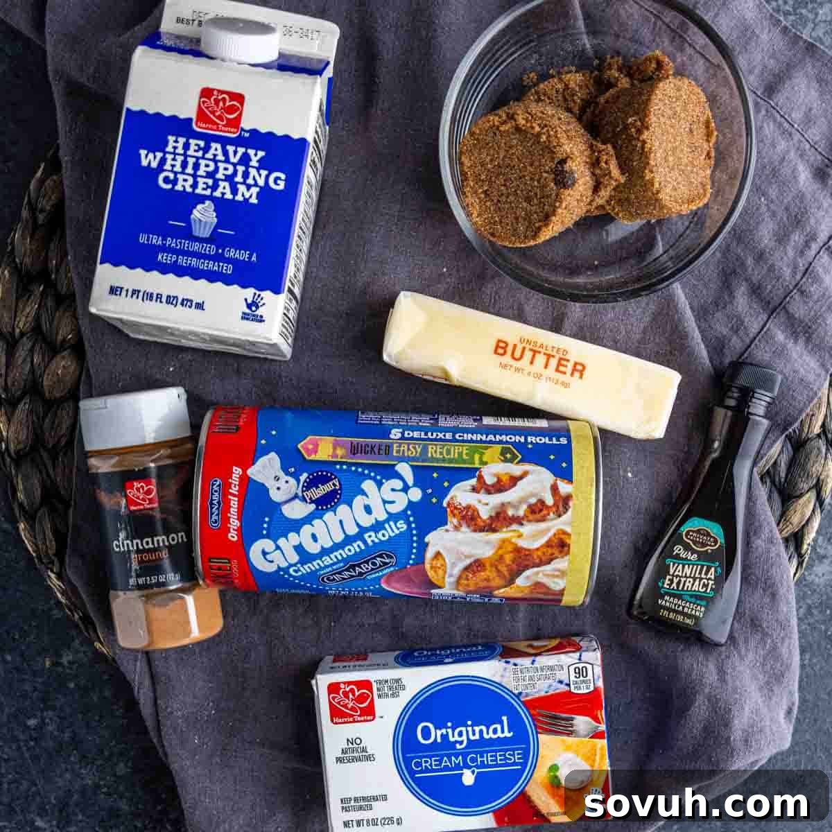 A beautifully arranged spread of ingredients for the viral TikTok cinnamon rolls with heavy cream. Featured prominently are a carton of heavy whipping cream, a bowl of rich brown sugar, a stick of golden butter, a sprinkle of aromatic ground cinnamon, a block of creamy cream cheese, a bottle of fragrant vanilla extract, and a fresh can of Pillsbury Grands! cinnamon rolls, all set on a rustic cloth.