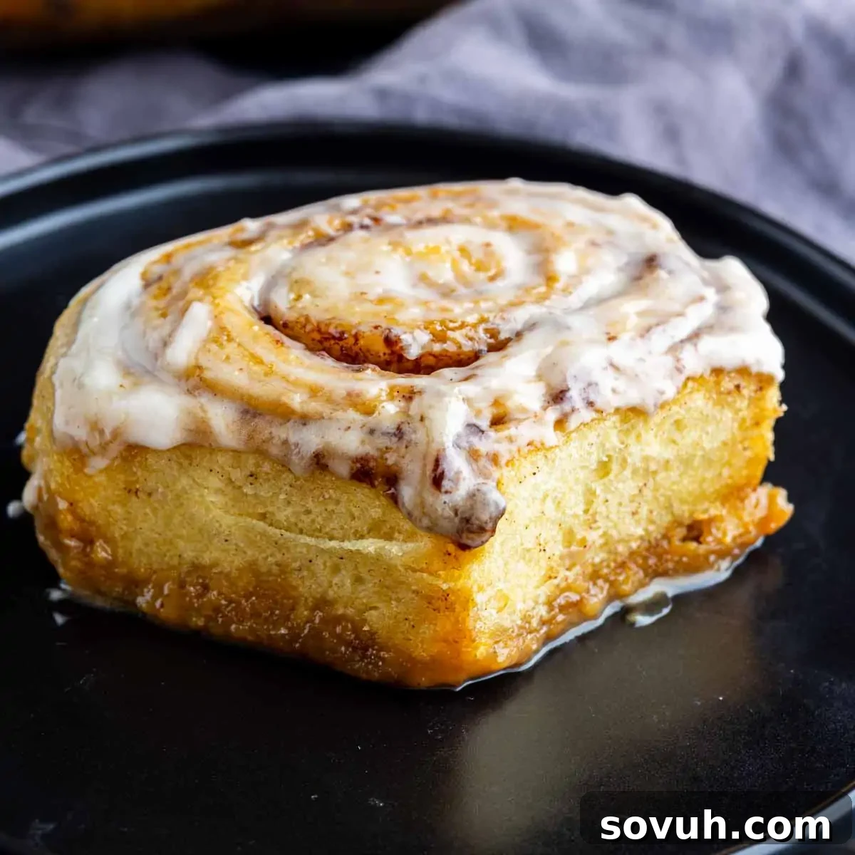 A perfectly baked and iced individual cinnamon roll sits on a sleek black plate, highlighting the effortless yet impactful transformation achieved by the viral TikTok cinnamon roll hack using heavy cream. The background is a soft gray cloth, allowing the roll to be the focal point.