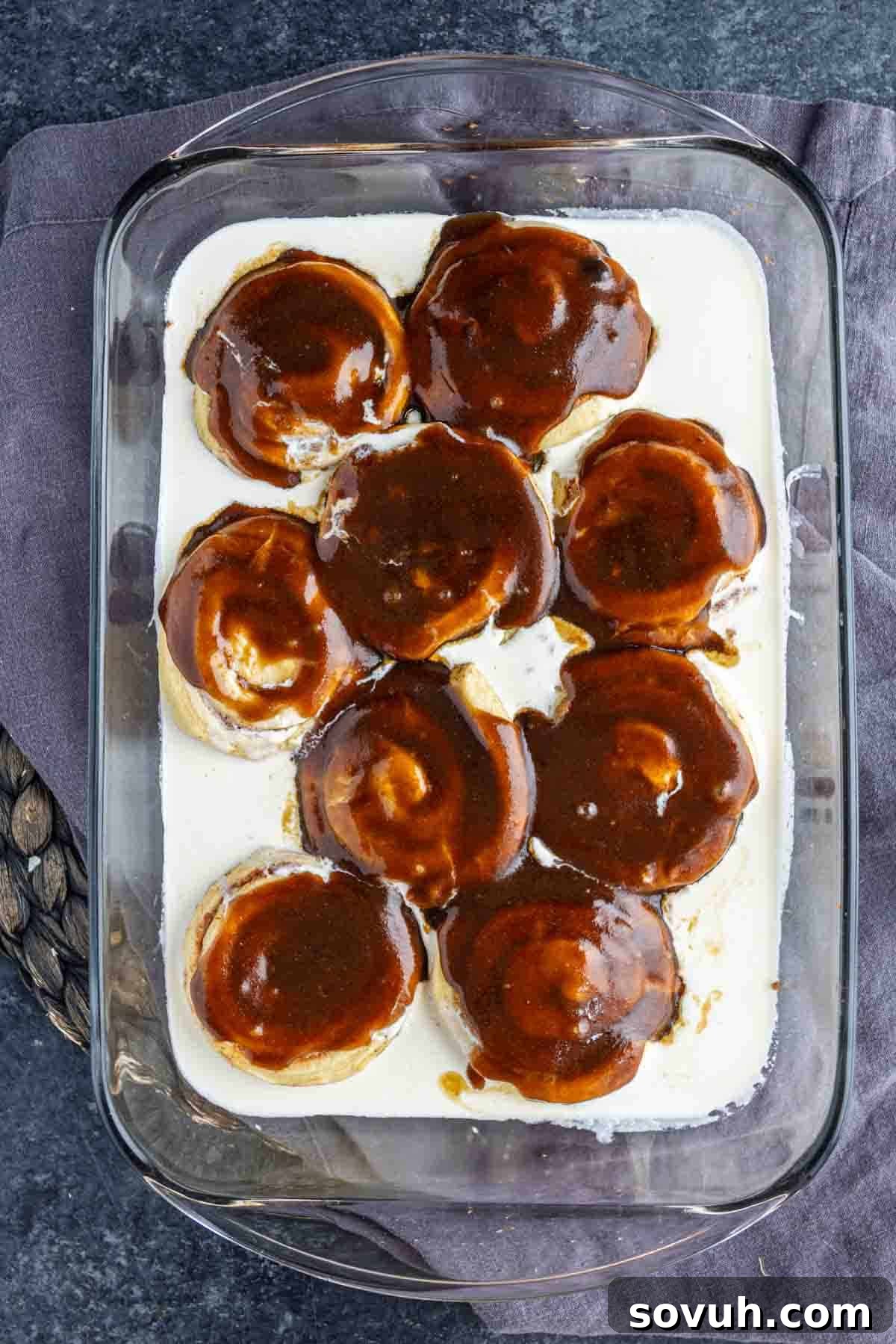 A glass baking dish showcasing eight unbaked cinnamon rolls bathed in heavy cream, generously covered with a rich brown sugar and cinnamon topping. This setup, on a dark surface with a purple napkin, illustrates the easy viral TikTok cinnamon roll hack before baking.