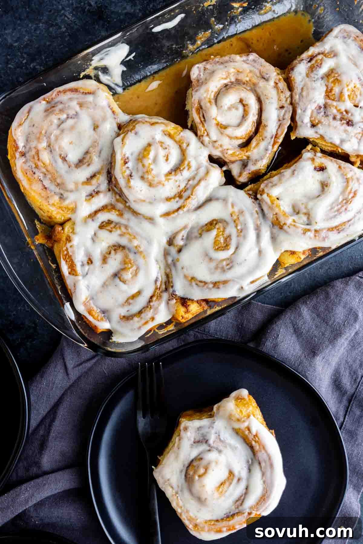 A tempting display of frosted TikTok cinnamon rolls with heavy cream in a glass baking dish, with one perfect, iced roll elegantly served on a black plate beside a fork. This composition emphasizes the ease and deliciousness of the viral TikTok recipe.