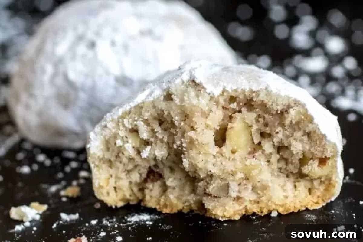 Close-up of a halved Mexican wedding cookie dusted with powdered sugar, revealing a crumbly interior with visible nuts. Another whole cookie is in the background.