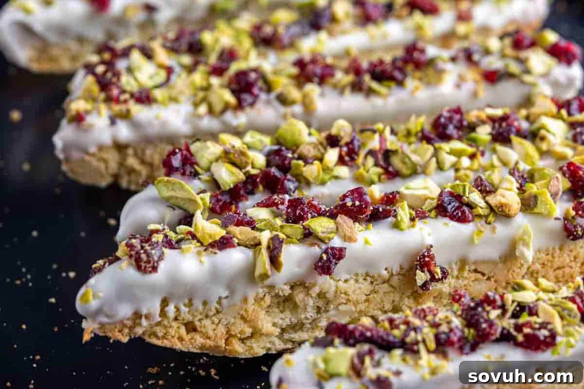 Biscotti topped with white chocolate, pistachios, and dried cranberries on a black surface, highlighting their crunchy texture.