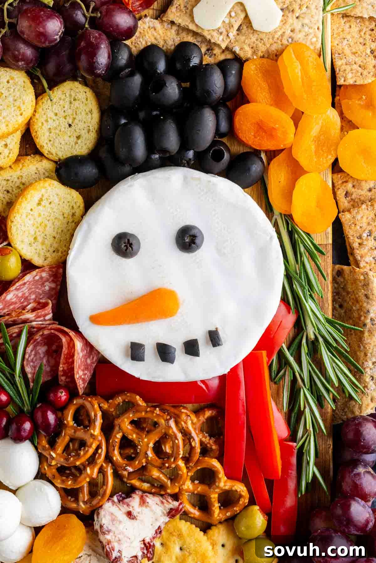 A cheese wheel decorated as a snowman face with olive hair, carrot nose, and olive eyes and mouth is the centerpiece of this festive holiday charcuterie board, surrounded by crackers, pretzels, dried apricots, grapes, herbs, and meats.