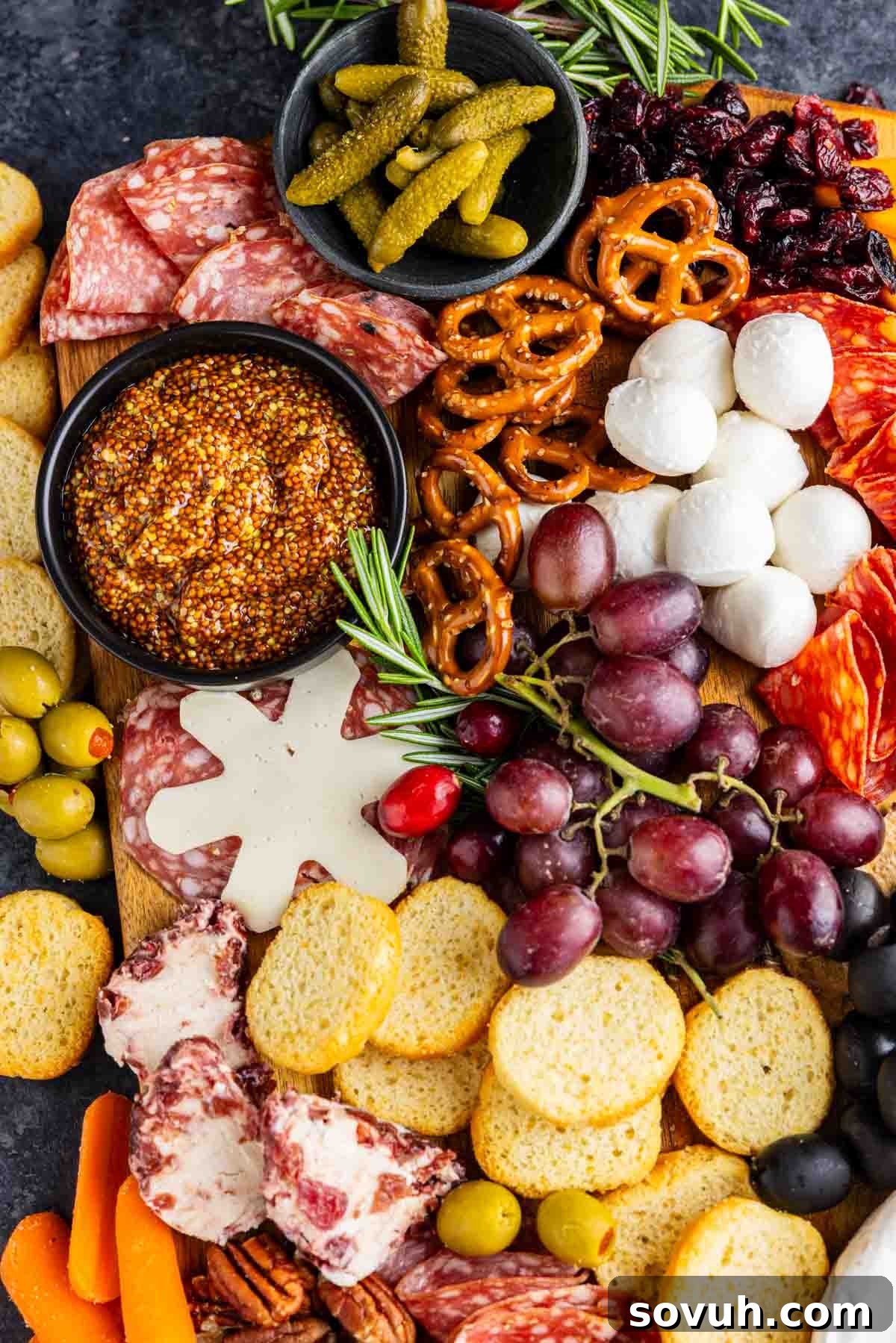 A festive charcuterie board with assorted meats, cheeses, crackers, grapes, olives, pretzels, pickles, mustard, dried cranberries, and sprigs of rosemary is perfect for easy holiday entertaining.