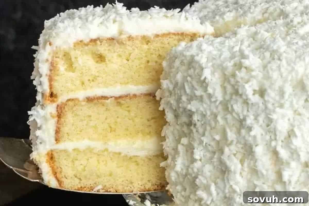 A three-layer vanilla cake with white frosting and shredded coconut on the outside, with one slice being served.