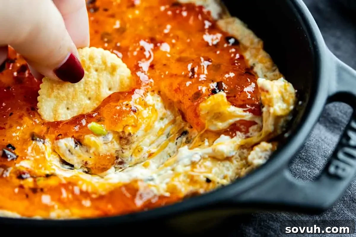 A hand dipping a round cracker into a creamy cheese dip topped with a layer of orange-red jelly in a black dish.