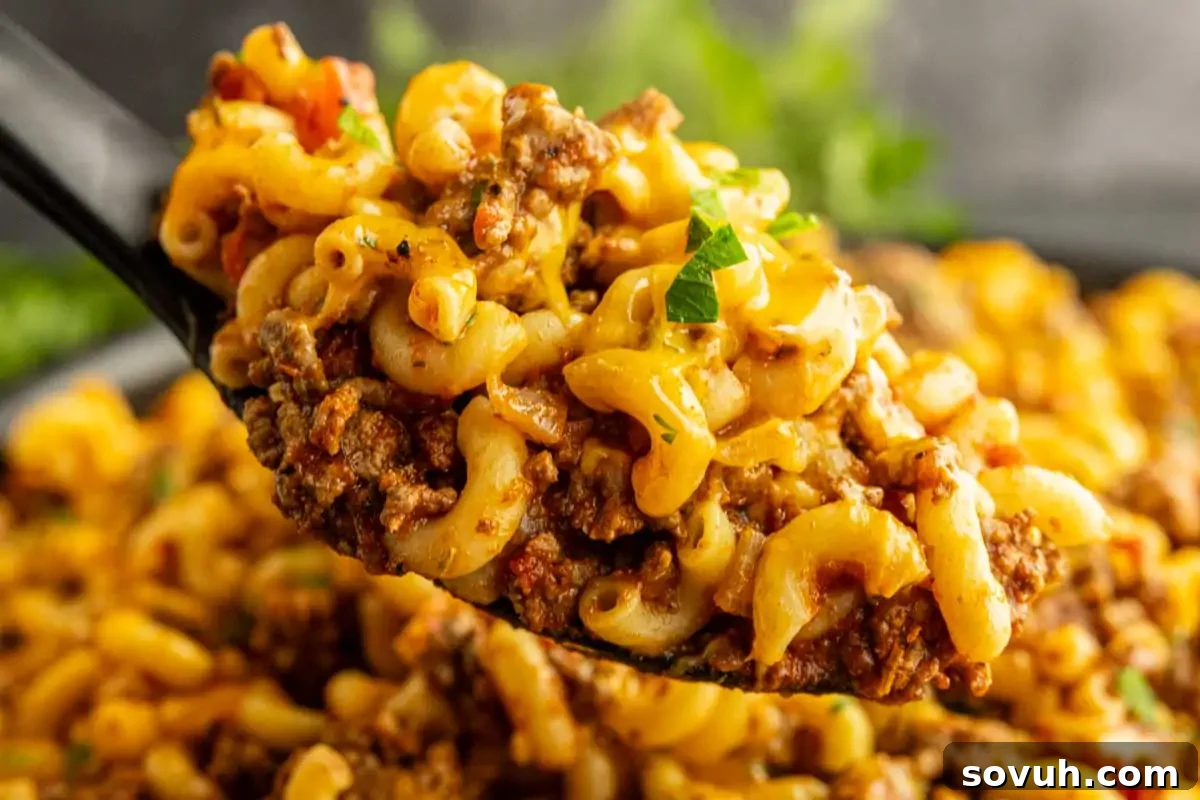 A close-up of a spoonful of macaroni pasta mixed with ground beef, tomato sauce, and melted cheese, garnished with chopped herbs.