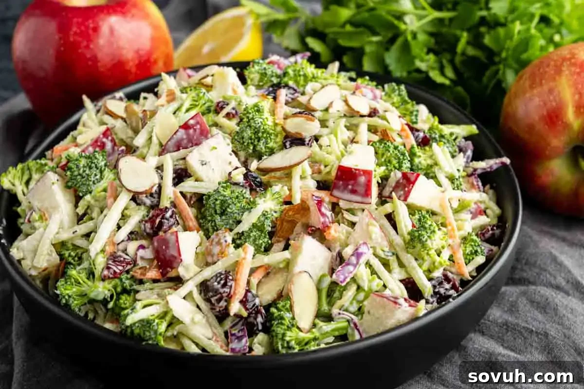 A bowl of salad with broccoli, apple chunks, shredded carrots, sliced almonds, and cranberries, with apples and herbs in the background.