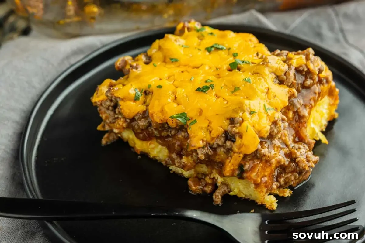 A slice of casserole topped with melted cheddar cheese and ground beef sits on a black plate with a black fork.