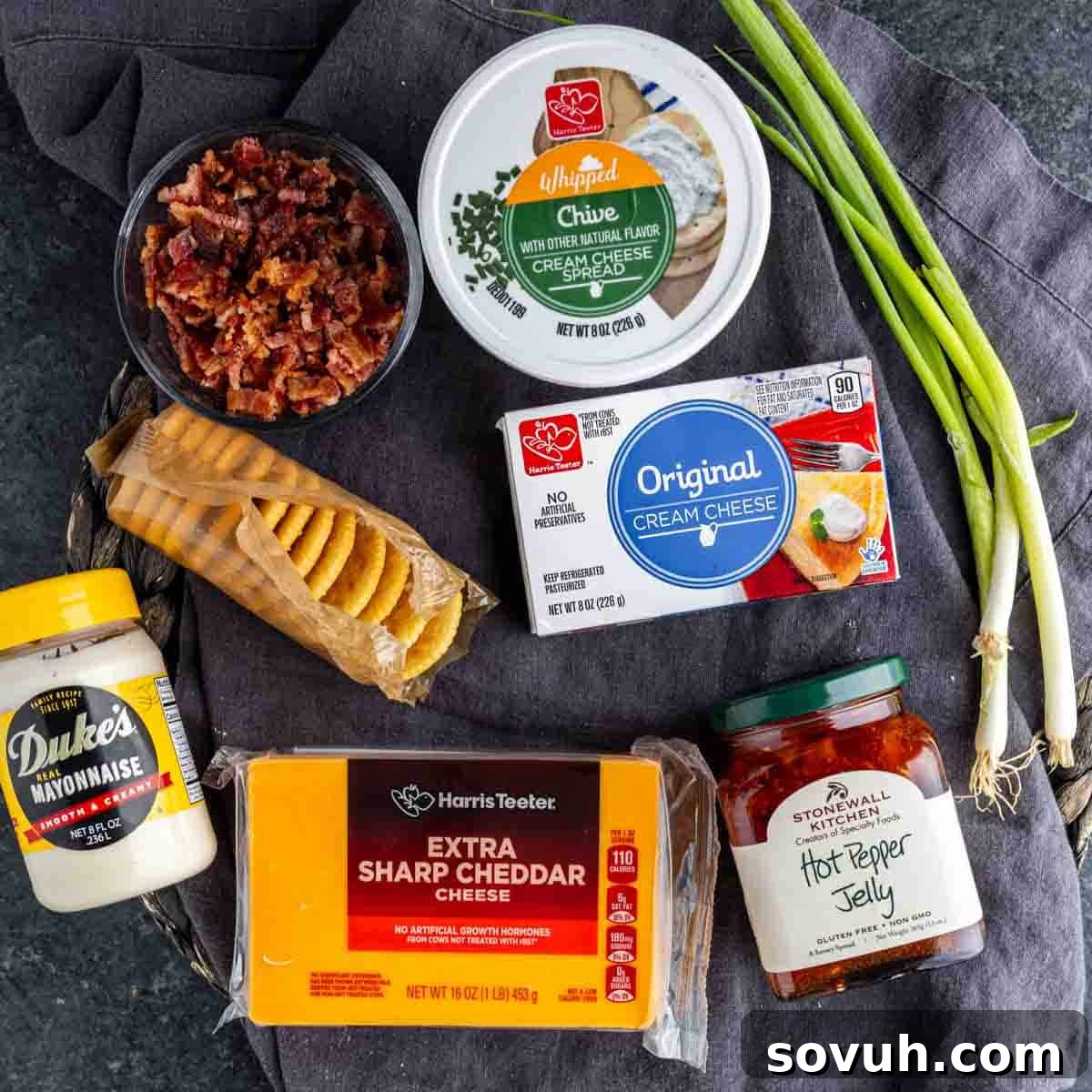 A flat lay of ingredients for a delicious hot pepper jelly appetizer, featuring bacon bits, whipped and original cream cheese, crackers, green onions, mayonnaise, sharp cheddar cheese, and hot pepper jelly on a dark cloth.