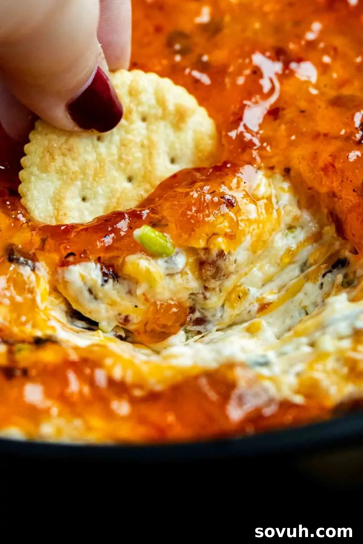 A hand dips a cracker into a hot, cheesy, baked cheese dip recipe with a bubbly golden topping—perfect for serving as a crowd-pleasing hot pepper jelly appetizer.