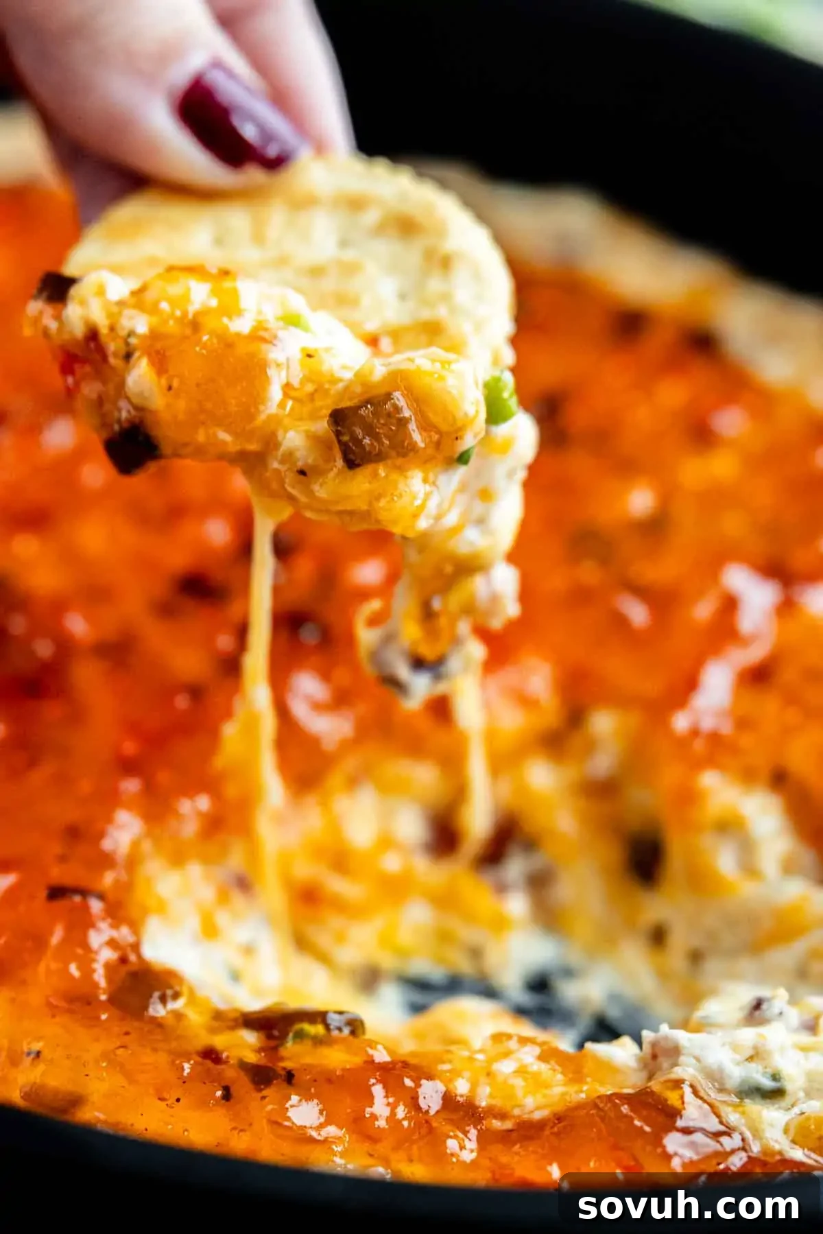 A hand holds a tortilla chip dipped into a cheesy, baked bacon and cream cheese dip, with melted cheese stretching from the chip to the dish.