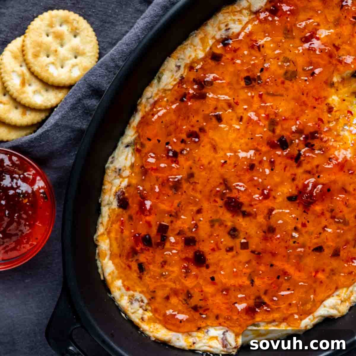 Baked cheese dip topped with hot pepper jelly in a black baking dish, served with round crackers and a small bowl of extra jelly on the side—a perfect hot pepper jelly appetizer for any gathering.