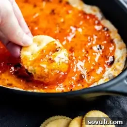 A hand dips a round cracker into a baking dish filled with a glossy, orange-colored spread or dip.