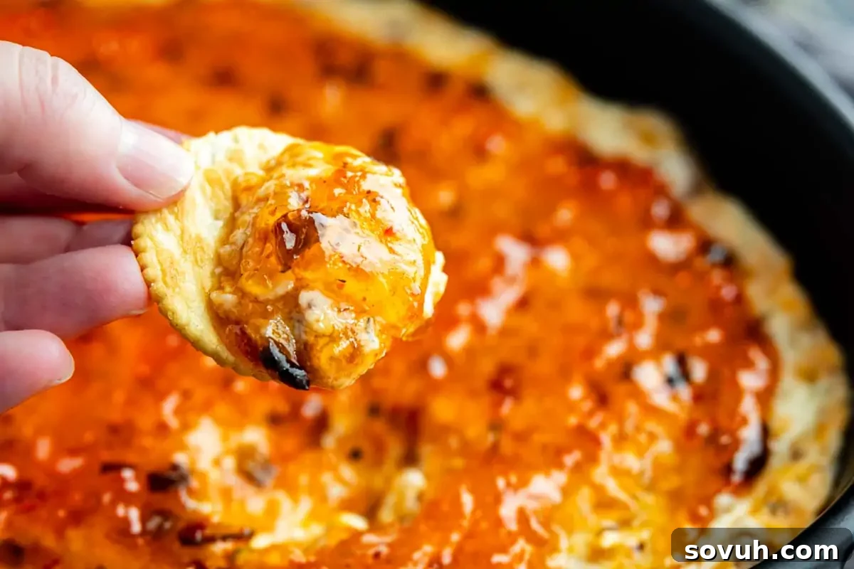 A hand holds a cracker piled high with a tantalizing hot pepper jelly dip, featuring creamy bacon and cream cheese. In the background, a warm pan is filled with the same savory appetizer, ready to be enjoyed at holiday gatherings or game day events.