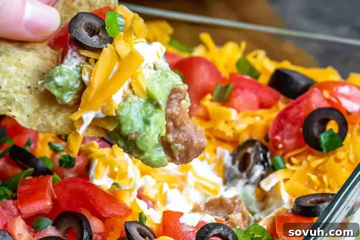 A vibrant close-up of a tortilla chip expertly scooping a generous portion of seven-layer dip, showcasing distinct layers of refried beans, creamy guacamole, rich sour cream, shredded cheese, diced tomatoes, sliced black olives, and fresh green onions, perfect for festive holiday parties.