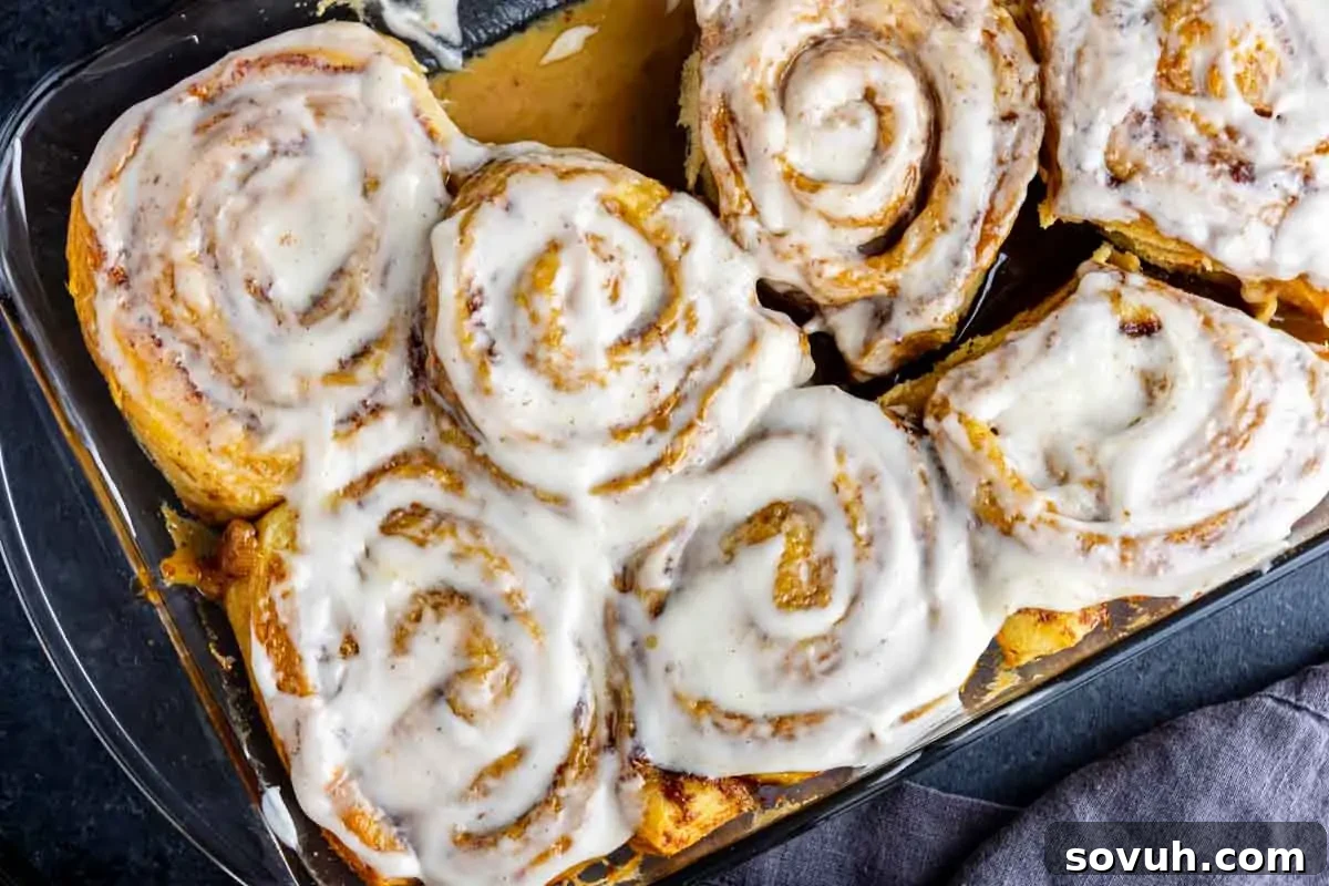 A glass baking dish filled with freshly baked cinnamon rolls covered in white icing, partially pulled apart, sitting on a dark surface.