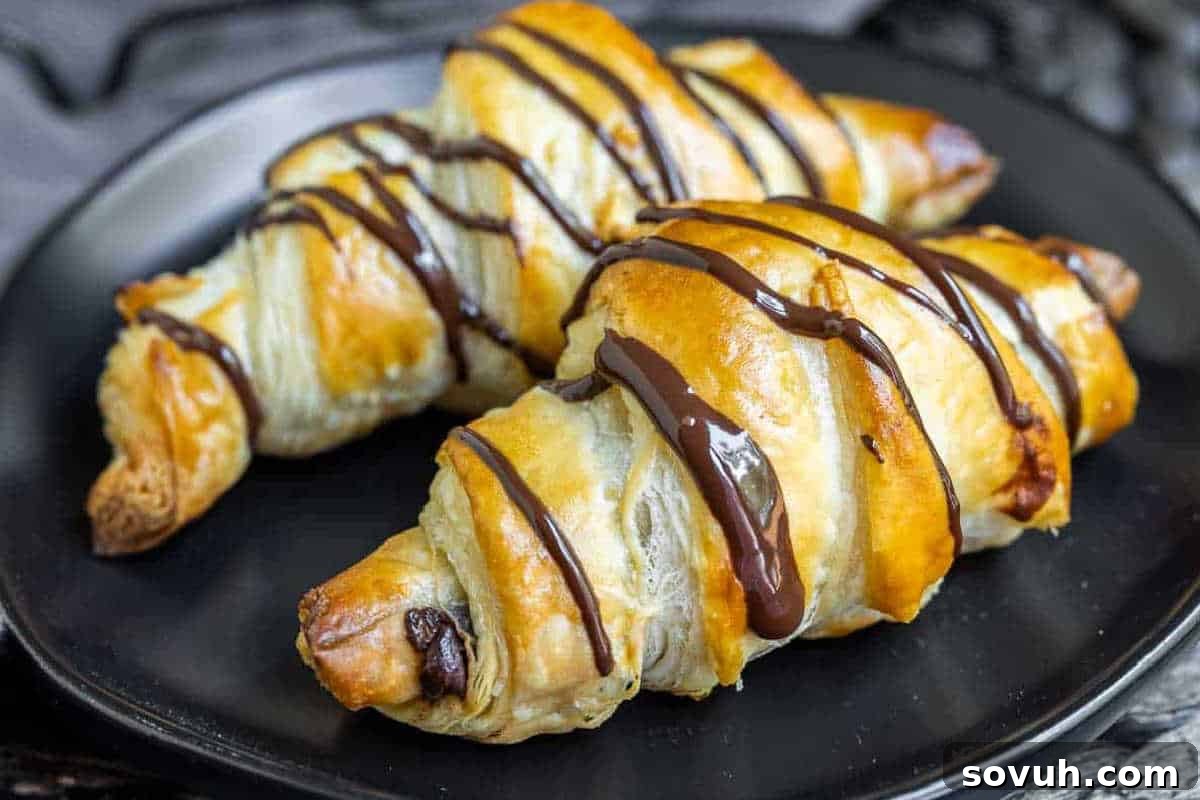 Two golden-brown croissants drizzled with chocolate on a black plate.