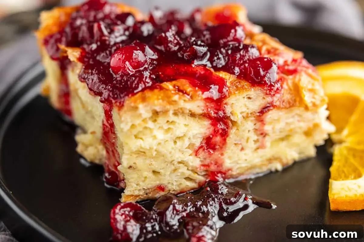 Close-up of a slice of bread pudding topped with a rich red berry sauce, served on a black plate with orange slices.