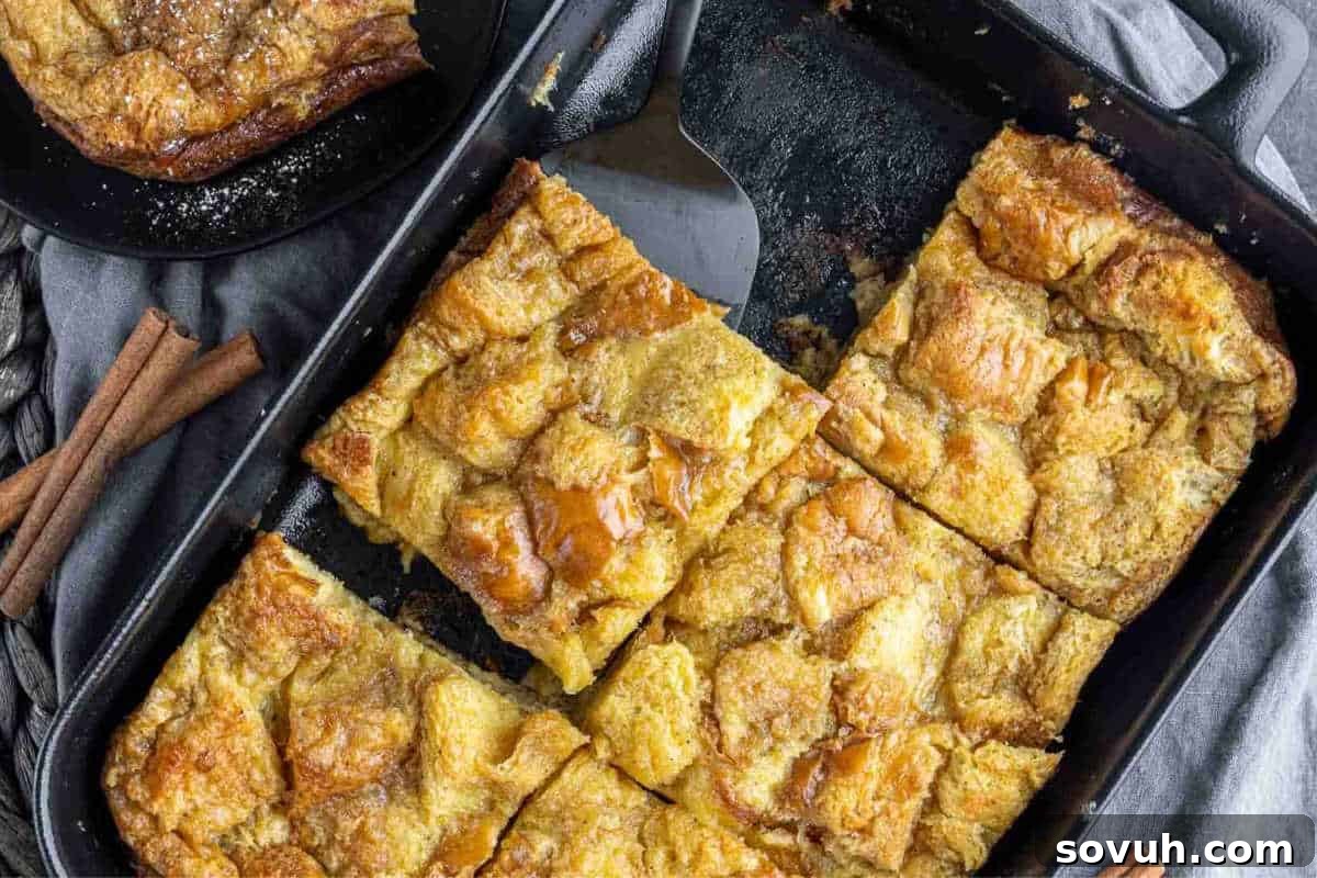A baking pan with four pieces of baked French toast topped with a glaze, next to a small plate with another piece. Cinnamon sticks are placed nearby on a gray cloth.