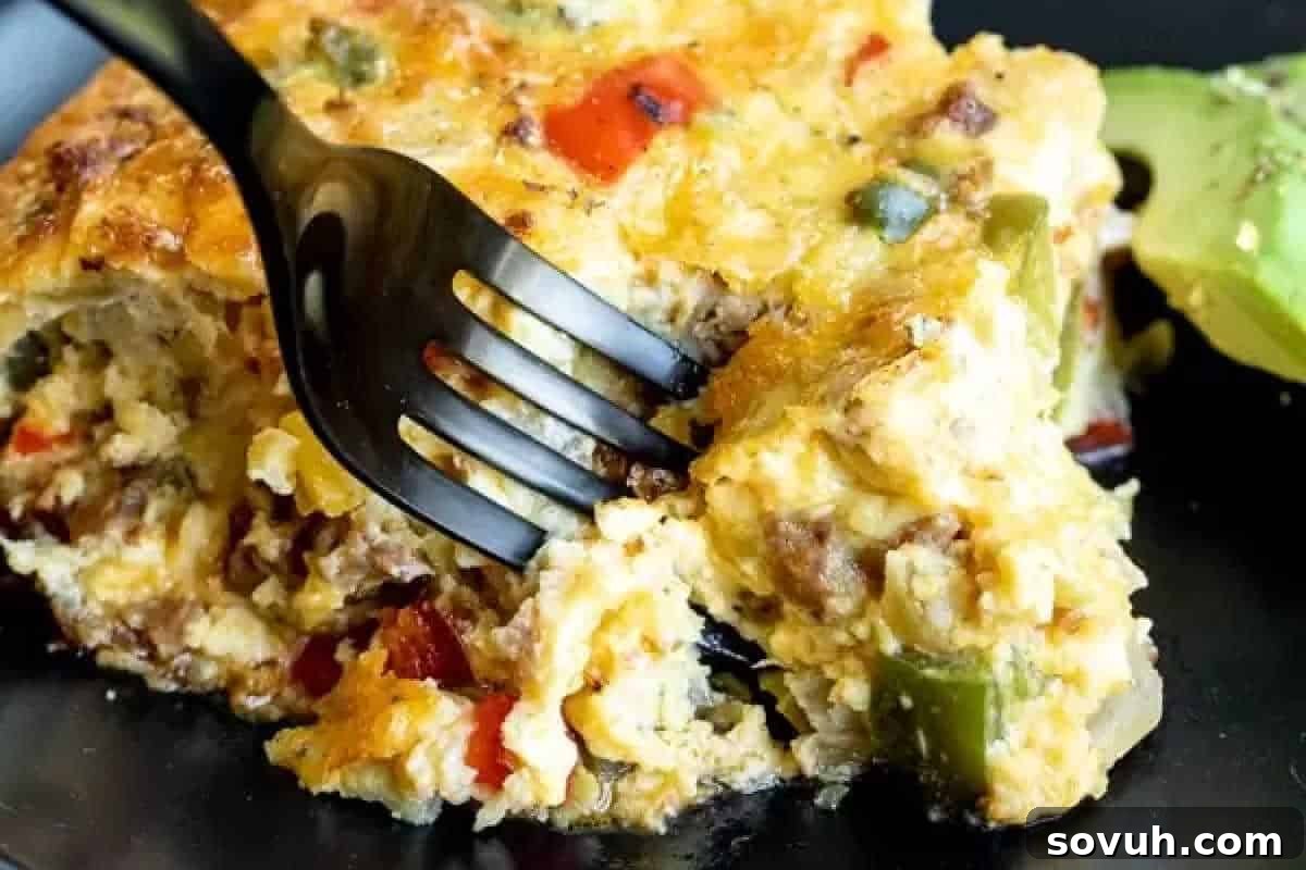 A close-up of a fork cutting into a slice of cheesy casserole with visible bell peppers and ground meat on a black plate. A slice of avocado is beside the casserole.