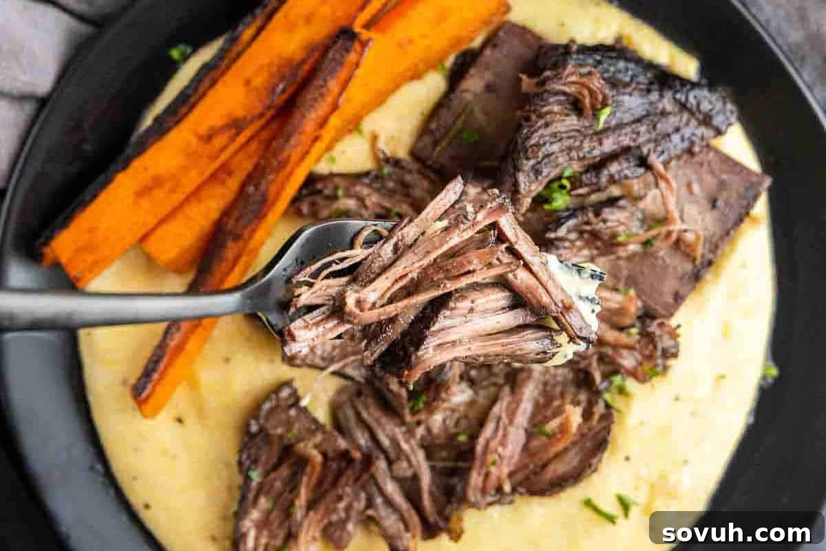 A fork lifts tender, shredded braised meat above a plate, showcasing a hearty meal of mashed potatoes, perfectly roasted carrots, and more succulent braised short ribs.