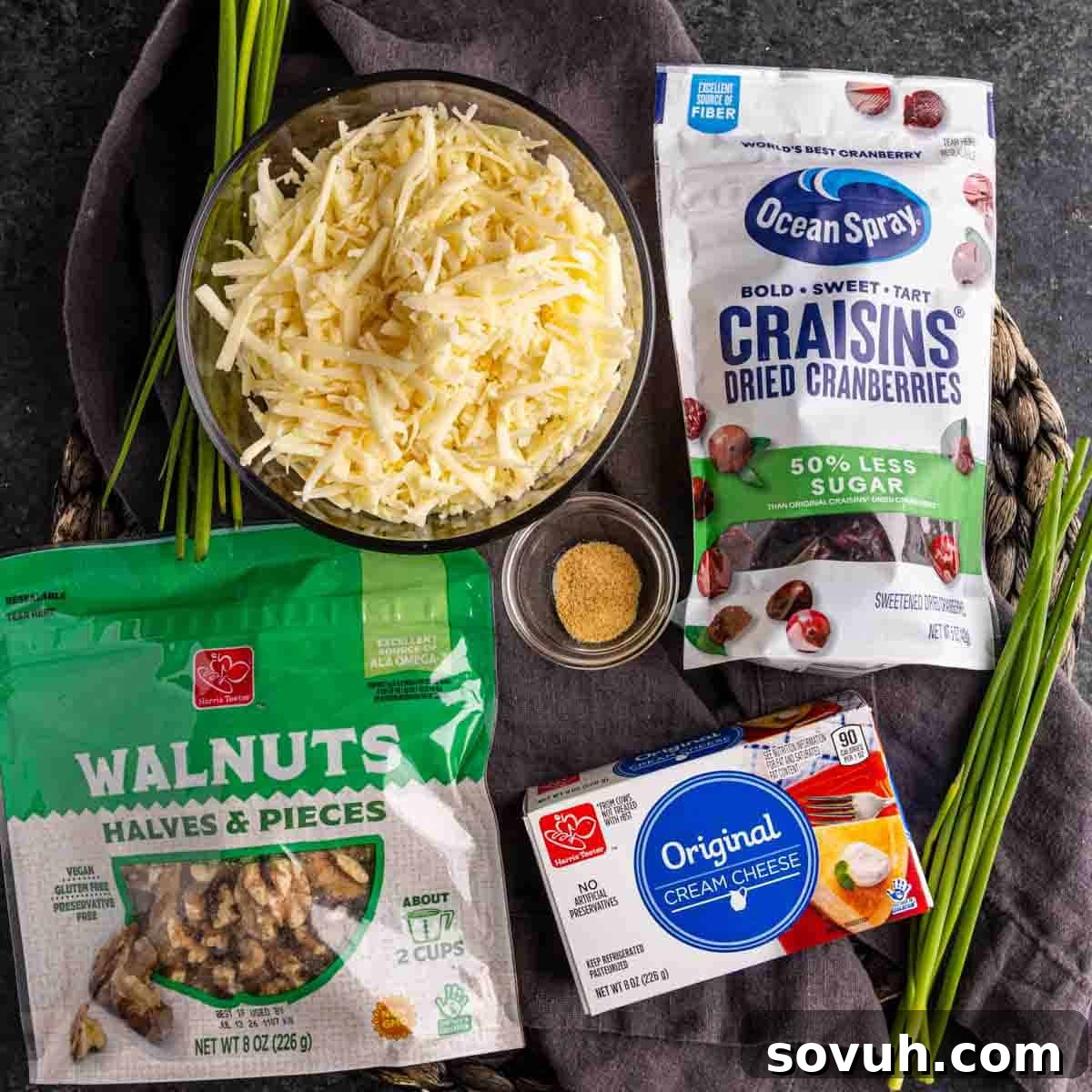 A vibrant collection of ingredients for an easy Cranberry Cheeseball recipe, featuring shredded white cheddar cheese, walnuts, dried cranberries, a block of softened cream cheese, savory seasoning, and fresh chives, ready to be transformed into a festive cheese ball with cranberries.