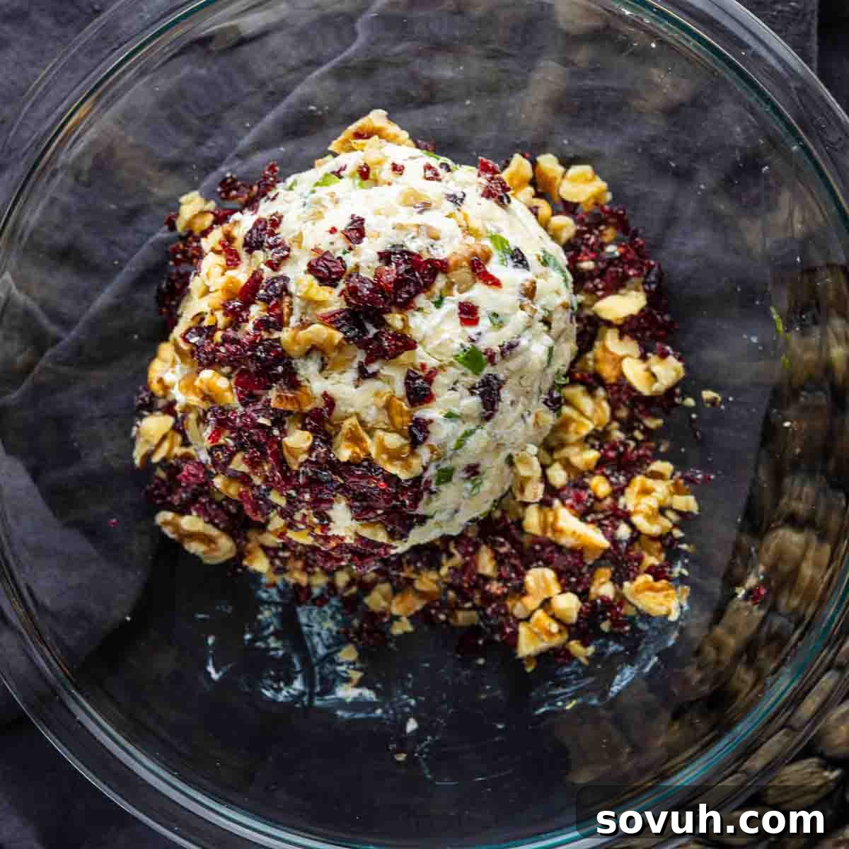A beautifully prepared Cranberry Cheeseball, generously coated with a vibrant mixture of chopped dried cranberries and walnuts, rests invitingly in a clear glass bowl on a dark cloth – a perfect festive appetizer.