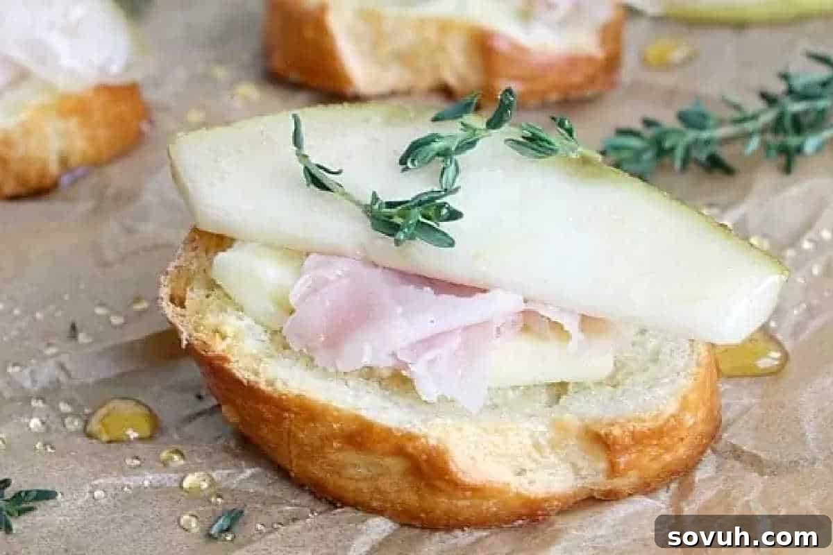 A gourmet appetizer featuring a crisp crostini slice, elegantly topped with tender pear, creamy Havarti cheese, savory ham, and a delicate sprig of fresh thyme, all beautifully drizzled with golden honey.
