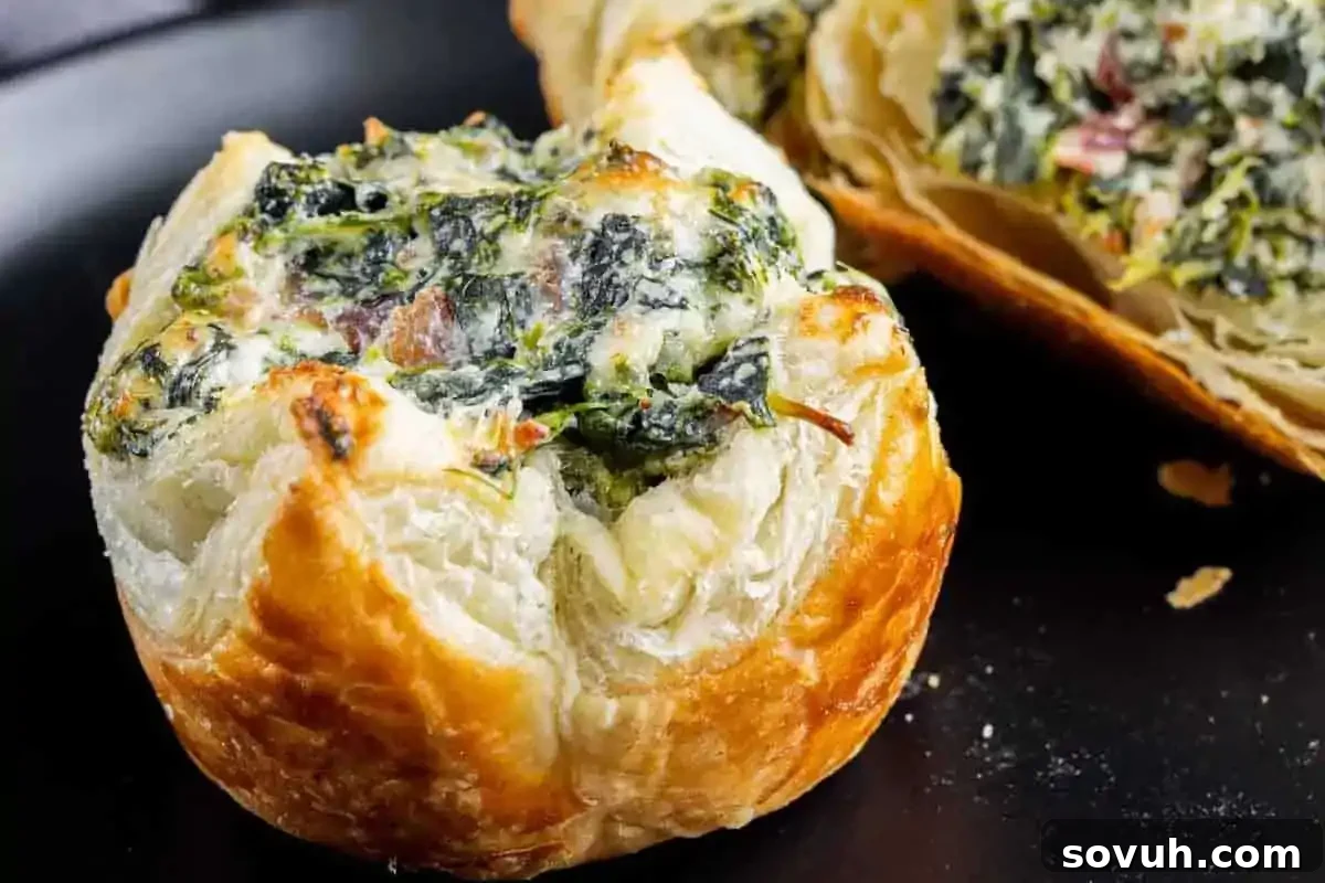A close-up view of a perfectly baked puff pastry, golden and flaky, generously filled with a creamy spinach and cheese mixture. A second pastry is artfully cut open, revealing its rich, inviting filling, ideal as elegant holiday appetizers or for a New Year’s Eve party spread.