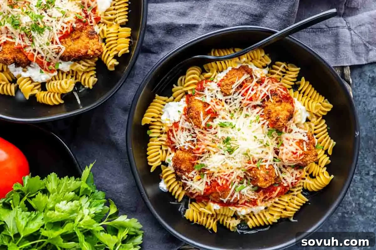 A vibrant black bowl filled with rotini pasta, rich tomato sauce, perfectly cooked breaded chicken pieces, dollops of creamy cottage cheese, and a sprinkle of grated Parmesan, garnished with fresh herbs. This easy high protein dinner is presented on a dark fabric background with fresh parsley and tomatoes nearby, highlighting its fresh appeal.