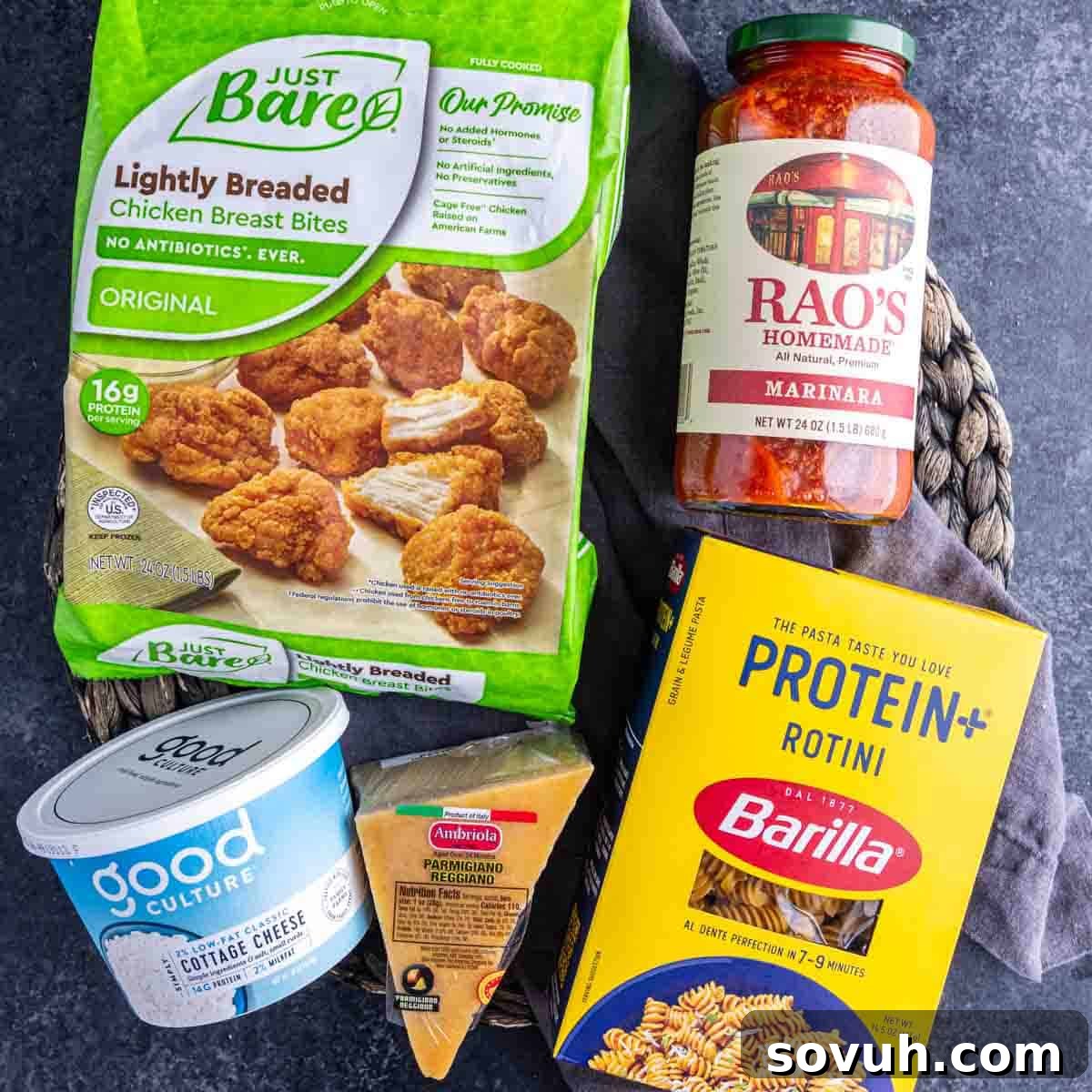 A selection of high-quality ingredients laid out: a bag of Just Bare chicken breast bites, a jar of Rao's marinara sauce, a box of Barilla Protein+ rotini pasta, a container of Good Culture cottage cheese, and a block of Parmesan cheese. These are all the essential components for creating an easy, high protein dinner or lunch.