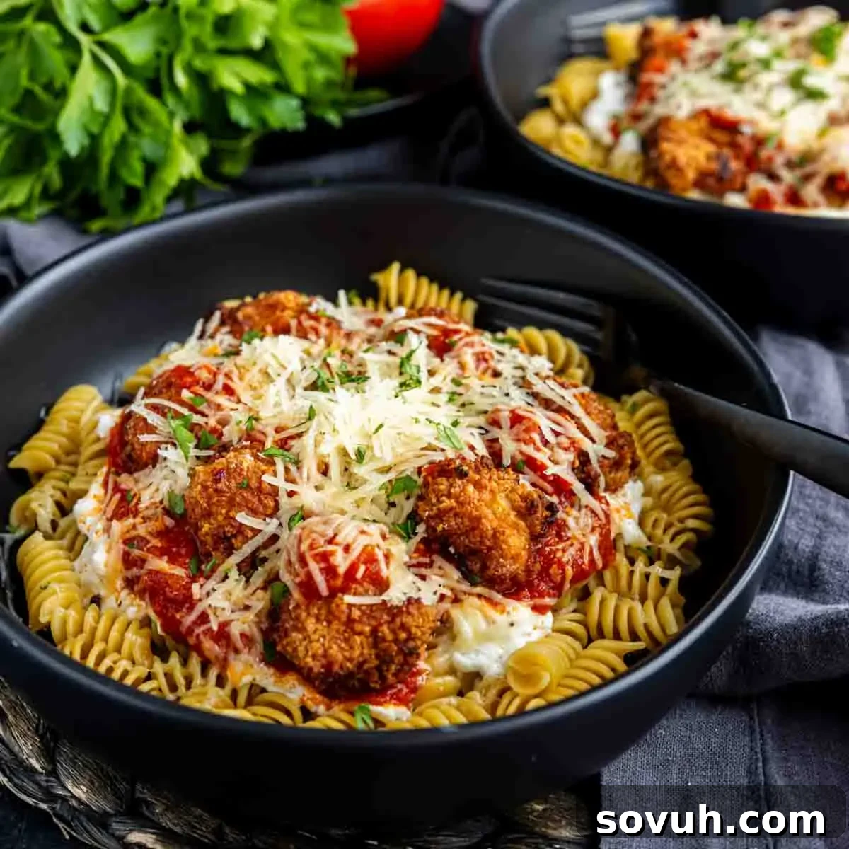 A close-up shot of a black bowl brimming with rotini pasta, topped with high-protein chicken pieces, generously coated in marinara sauce, and finished with a cascade of grated cheese and fresh herbs. A fork is casually placed in the bowl, ready to dig in, and fresh parsley sprigs are arranged nearby. This image highlights an easy, high-protein lunch option that's both appealing and nutritious.