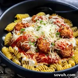 A black bowl filled with rotini pasta topped with meatballs, marinara sauce, grated cheese, and chopped herbs, with a black fork on the side.