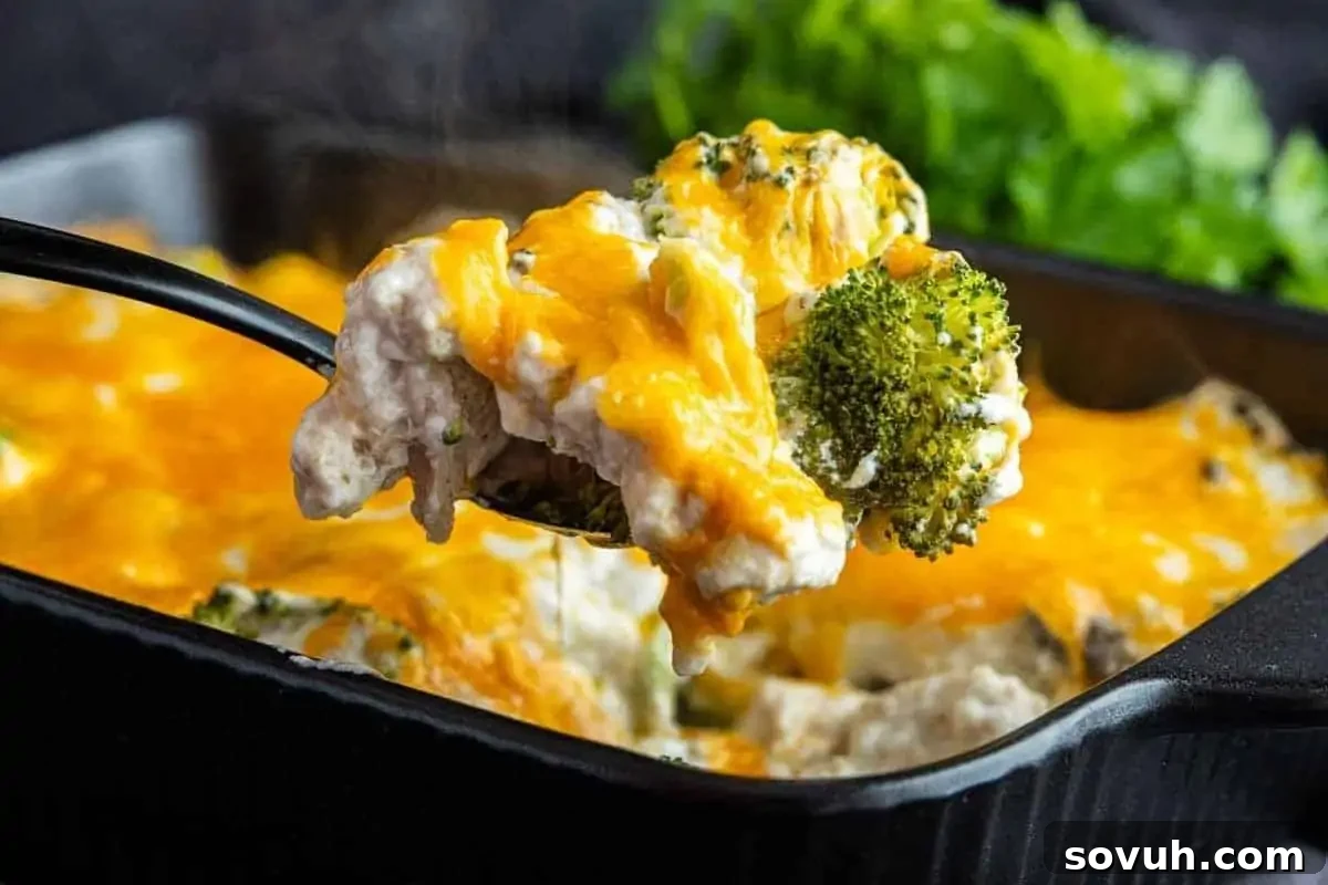 A spoon lifts a serving of cheesy broccoli casserole from a black baking dish, with melted cheese and broccoli visible, and fresh herbs in the background. This is a perfect high-protein, low-calorie, healthy meal option.