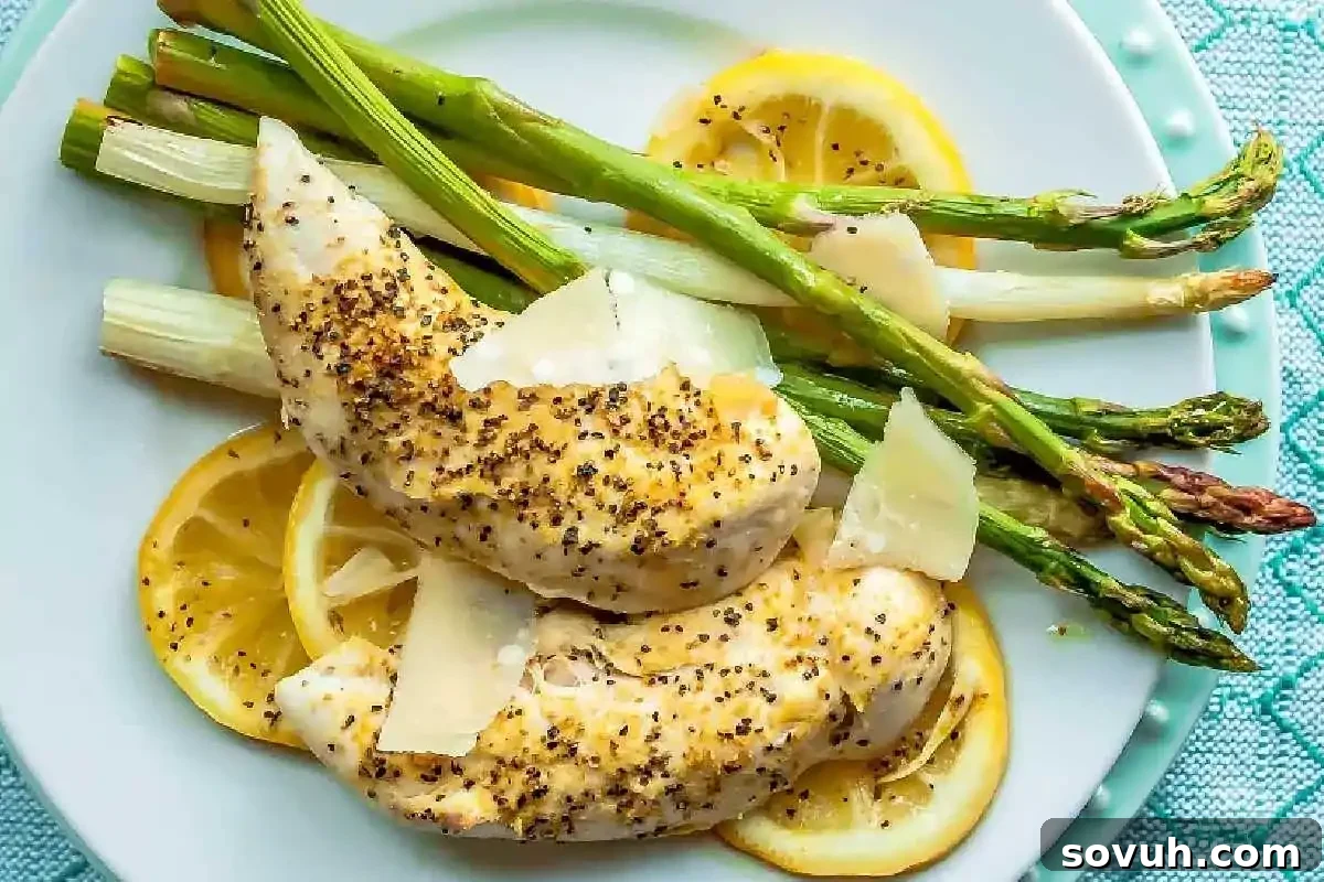Two pieces of seasoned baked chicken breast served on lemon slices with steamed asparagus and shaved parmesan—a delicious option for best high-protein recipes to start your new year recipes collection. A quick and flavorful sheet pan lemon chicken and asparagus dinner.