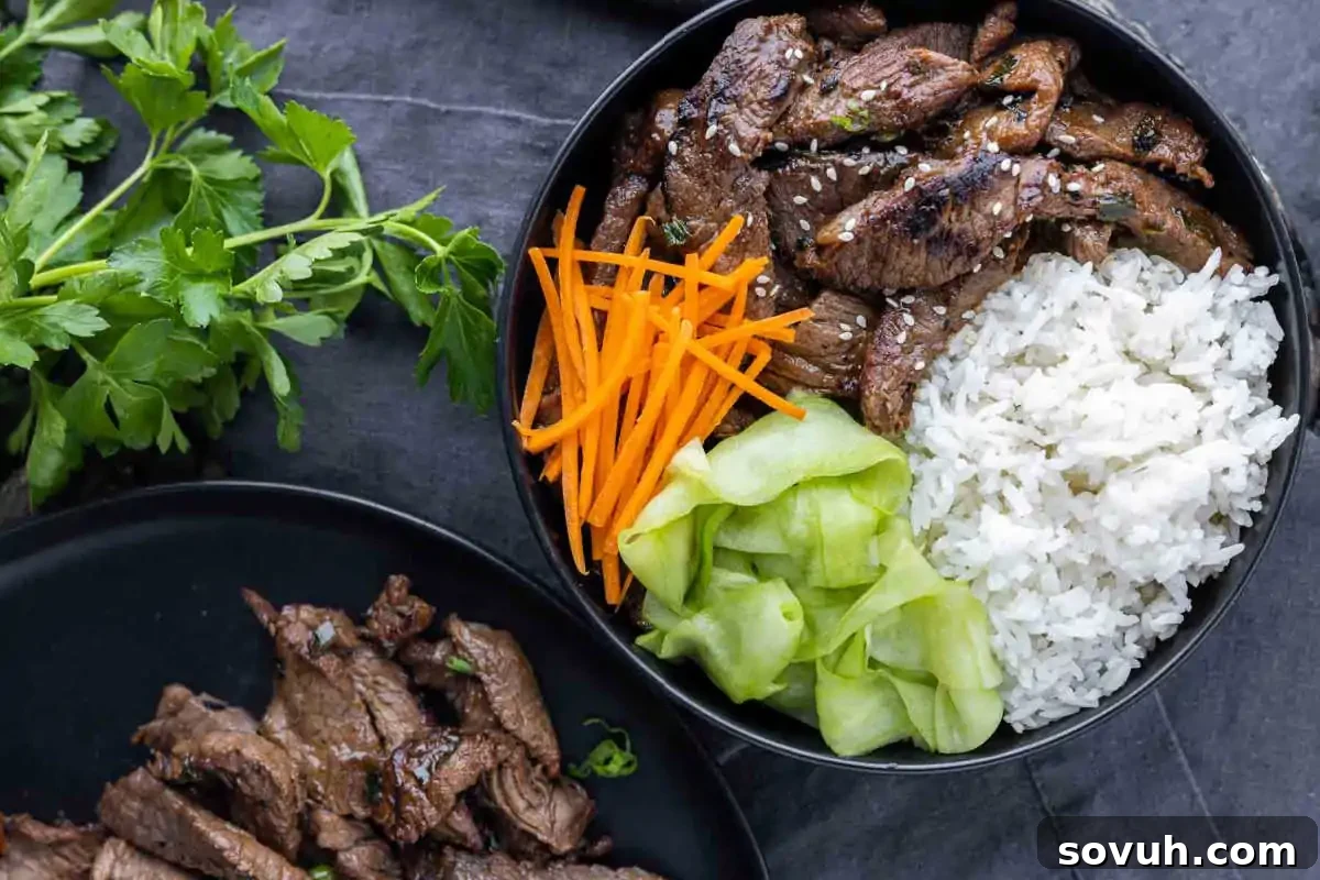 A bowl with sliced grilled beef, white rice, shredded carrots, and cucumber ribbons, garnished with sesame seeds, sits next to a plate of additional beef—one of the best recipes for high-protein meals to start the New Year right. A delicious and savory Korean beef bulgogi dish.