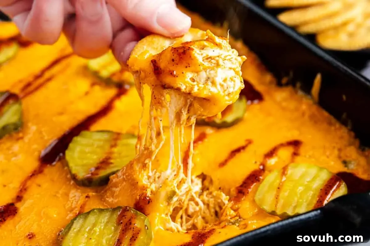 A person holding a cracker with melted cheese over a Nashville Hot Chicken Dip, garnished with pickles, ready for serving as a game day dip or party appetizer.