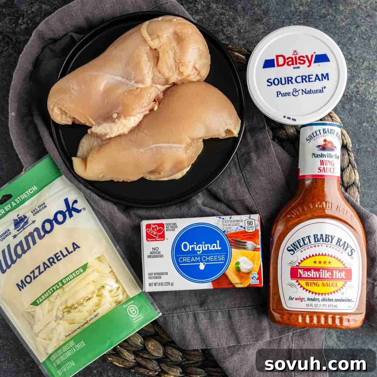 Raw chicken breasts on a black plate, alongside packages of mozzarella cheese, cream cheese, sour cream, and a bottle of Nashville hot wing sauce – essential ingredients for crafting the perfect hot chicken dip or party dip.