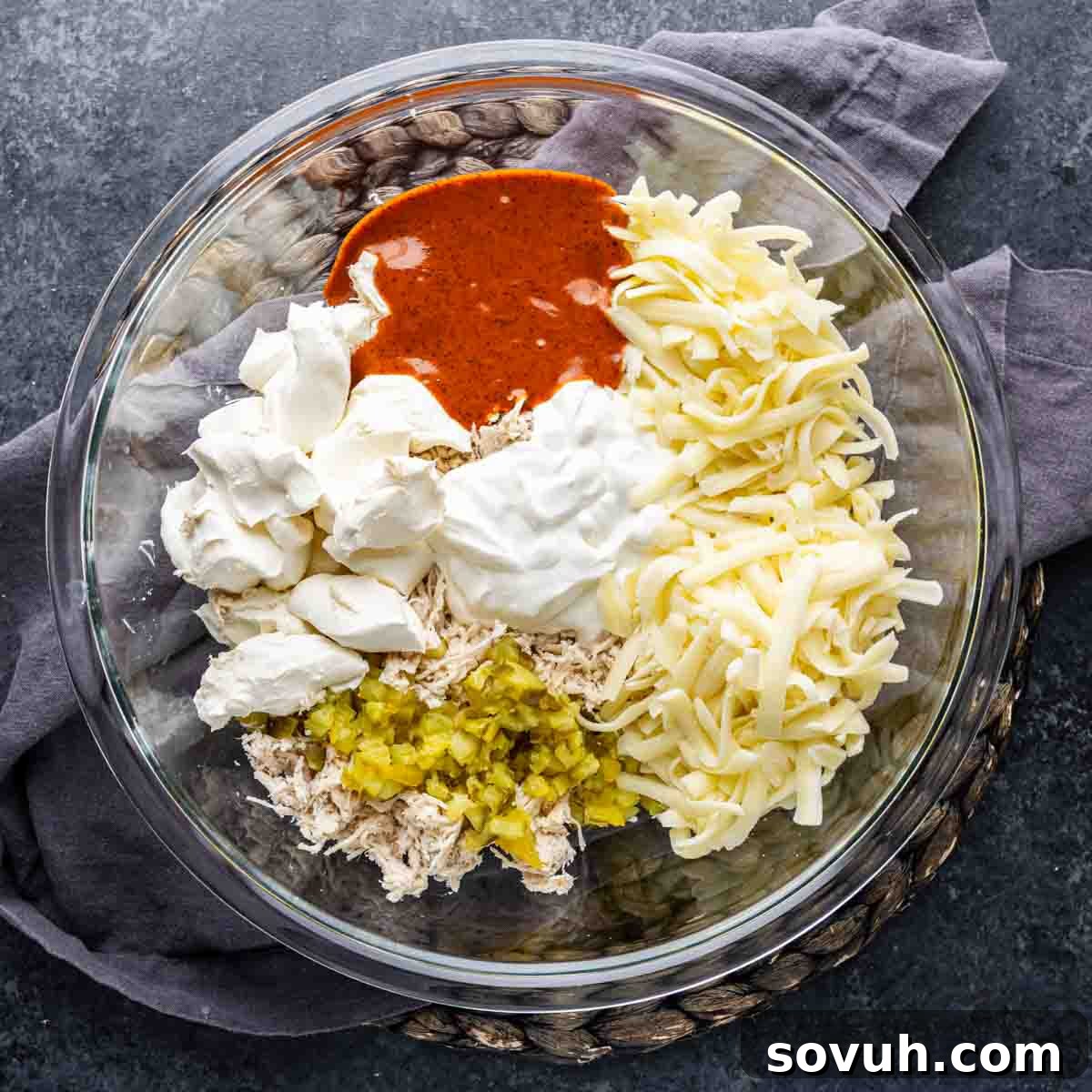 A glass bowl with shredded chicken, chopped pickles, shredded cheese, sour cream, cream cheese, and a reddish sauce—all the essential components for a game day dip—arranged on a gray napkin atop a dark surface.
