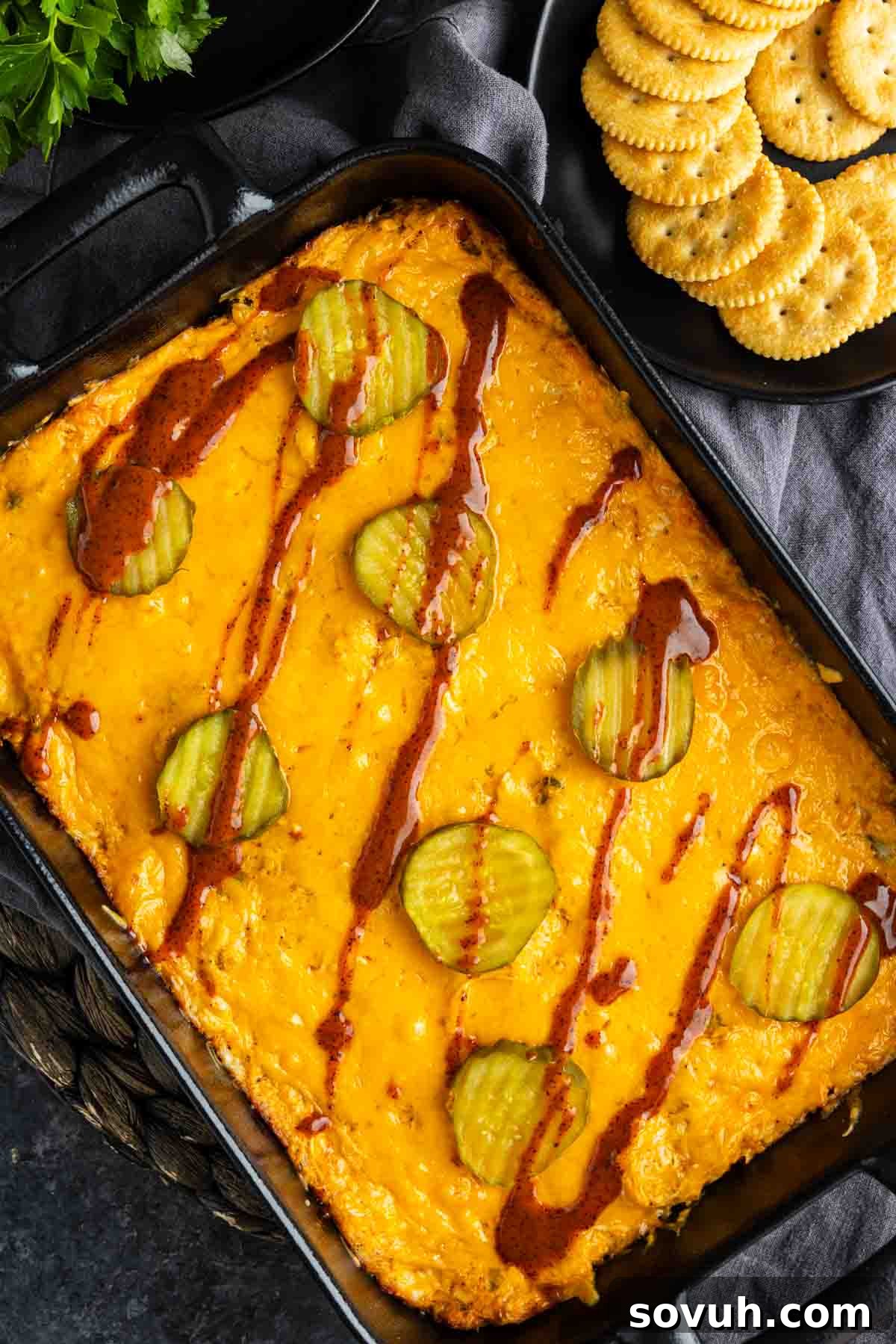 Rectangular baking dish featuring Nashville hot chicken dip with melted cheese, topped with pickle slices and a drizzle of sauce. A plate of round crackers is visible in the upper right corner, ready for serving.