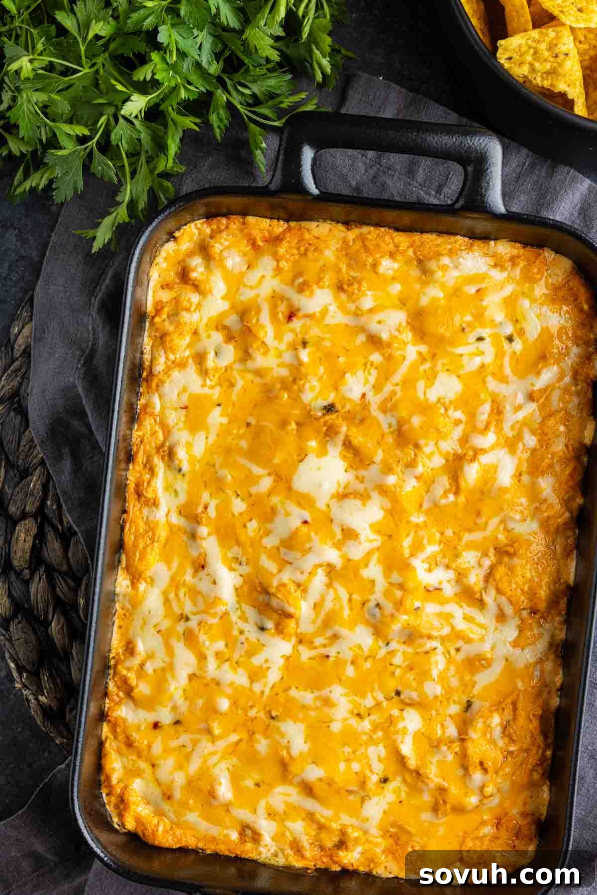 A baking dish filled with golden, melted cheese-topped chicken enchilada dip sits on a dark surface next to fresh parsley and a bowl of tortilla chips.