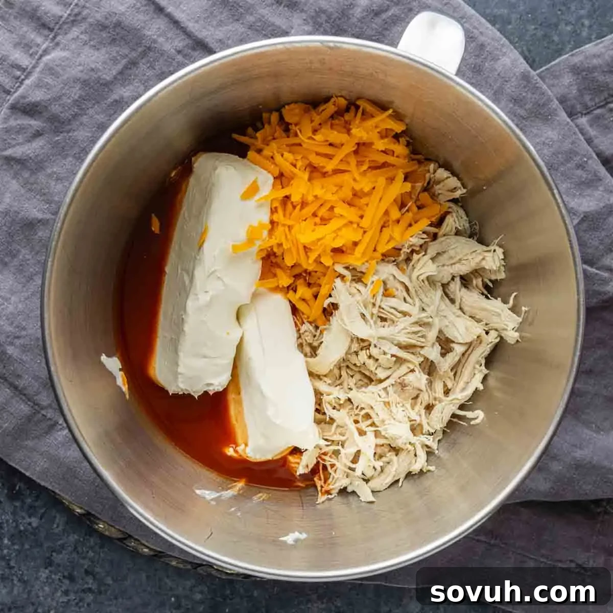 A metal mixing bowl containing shredded chicken, cheddar cheese, cream cheese blocks, and buffalo sauce sits on a gray cloth—perfect for a zesty baked chicken dip or as a crowd-pleasing game day dip.