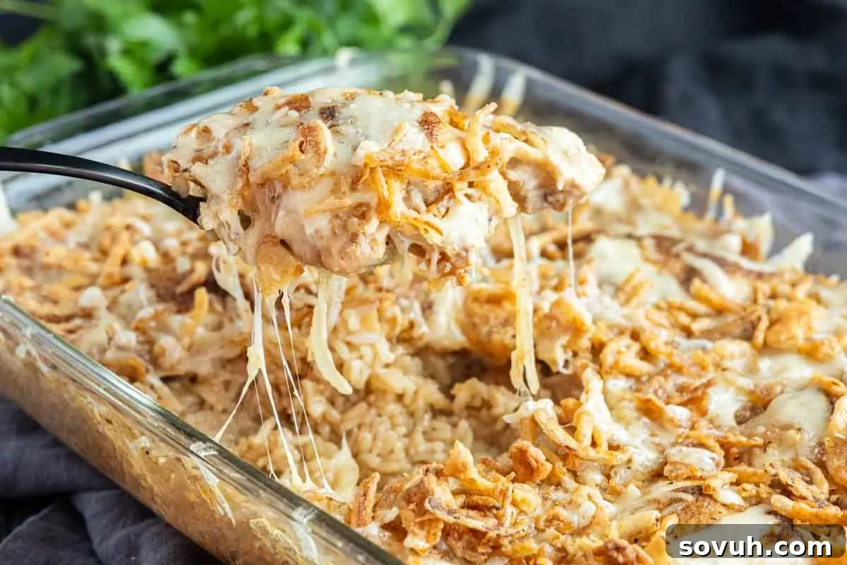A serving spoon lifts a portion of cheesy, baked French Onion Chicken Bake with crispy fried onions from a glass dish.