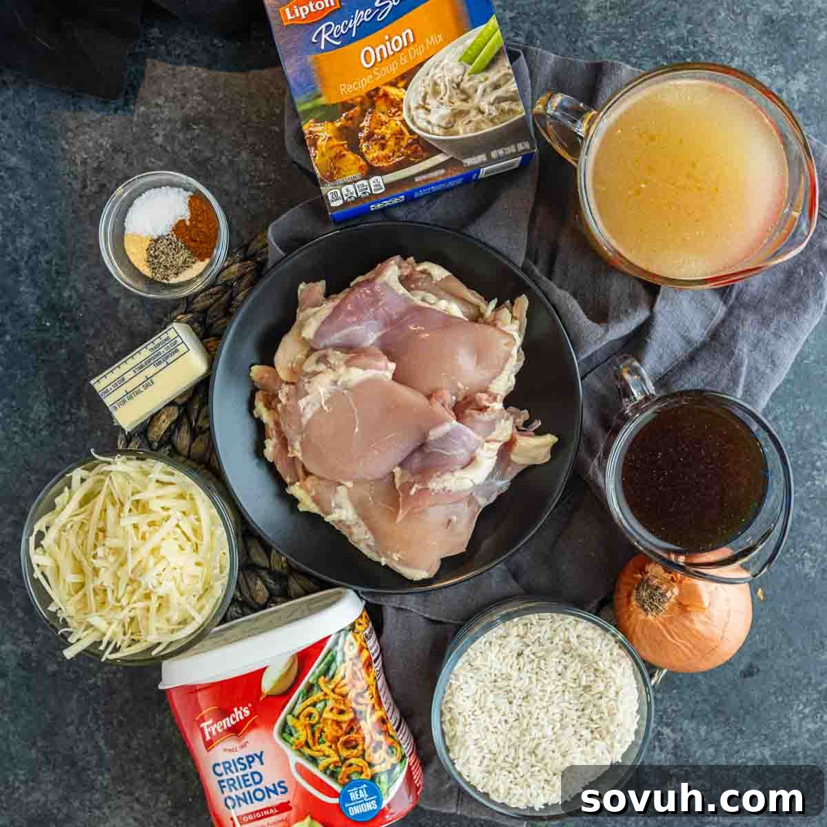A flat lay of ingredients for a French Onion Chicken and Rice Bake, including raw chicken thighs, cheese, rice, onion, broth, soup mix, seasonings, butter, crispy fried onions, and soy sauce.