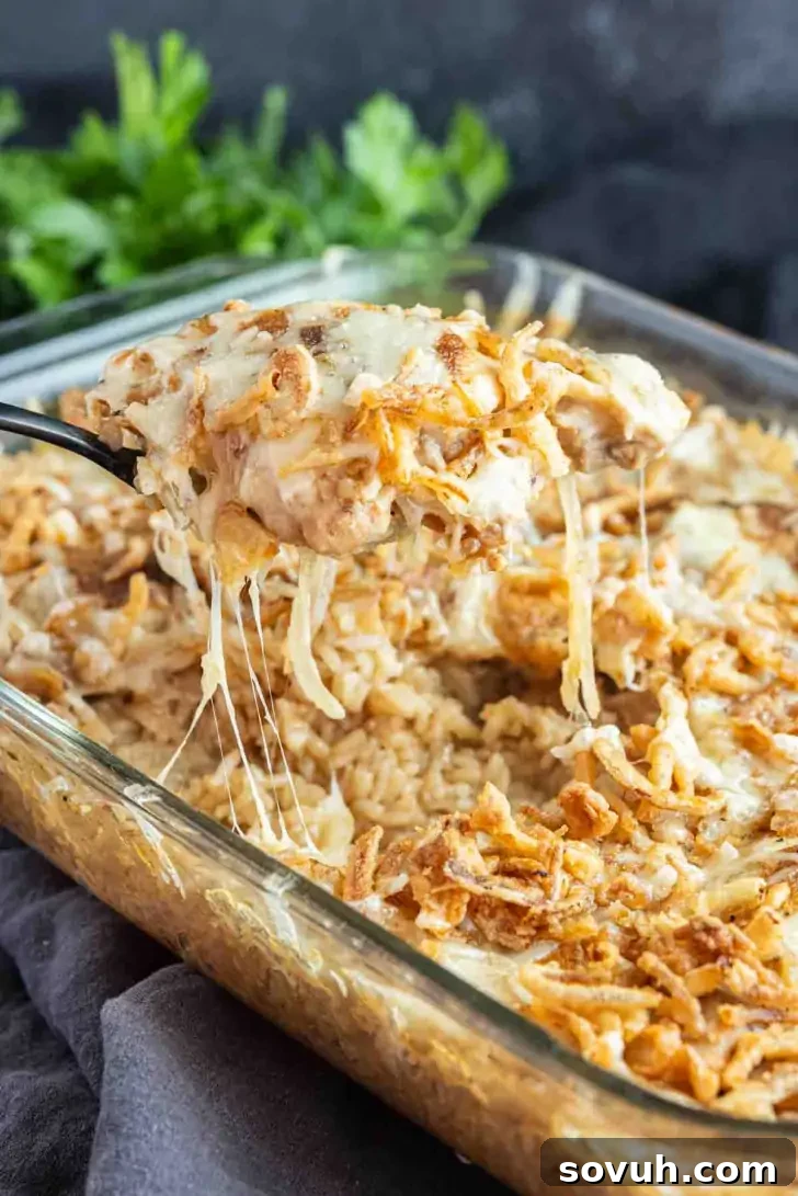 A casserole dish filled with a creamy, cheesy French Onion Chicken and Rice Bake topped with crispy fried onions; a serving is being lifted with a spoon.