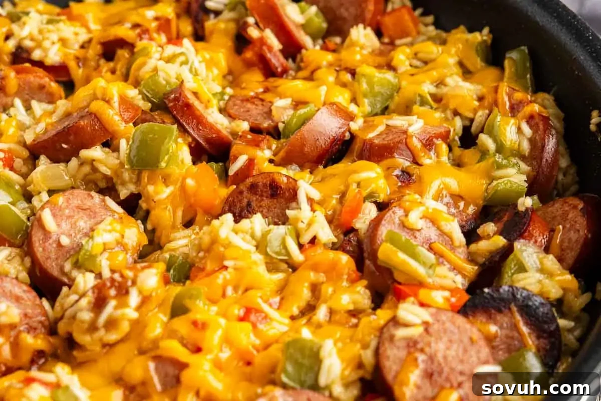 A close-up of a skillet containing sliced sausage, rice, diced green peppers, and melted cheddar cheese—an irresistible one pot cheesy sausage and rice dish.