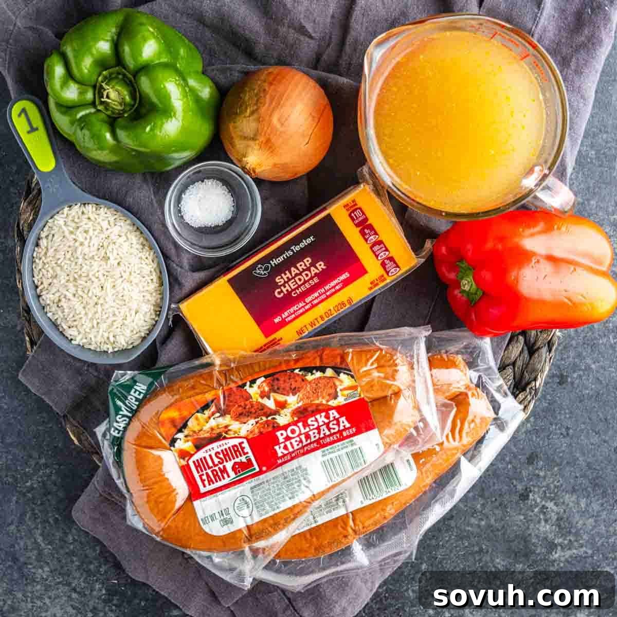 Ingredients for one pot cheesy sausage and rice on a dark surface: green bell pepper, yellow onion, salt, white rice, cheddar cheese, chicken broth, red bell pepper, and Polska kielbasa sausage.
