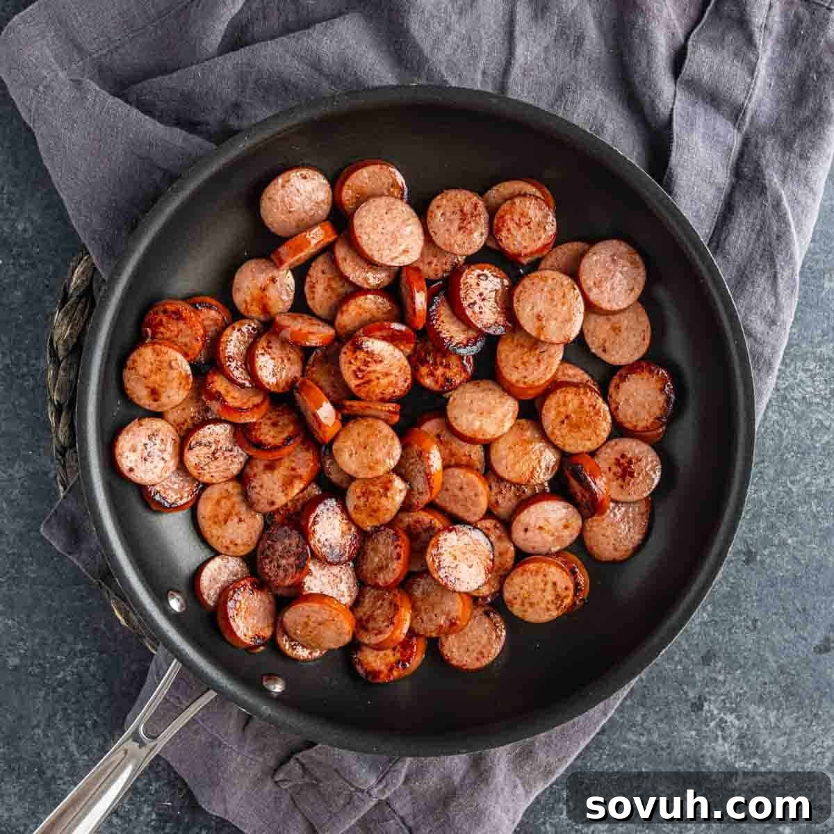 Sliced sausage pieces being browned in a black frying pan, perfect for starting a cheesy sausage and rice dish, with the pan resting on a gray cloth.